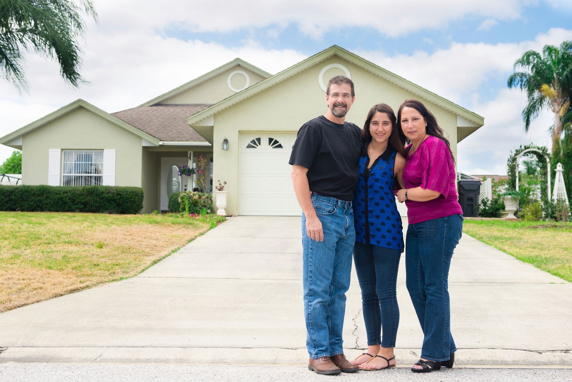 A family of three standing on a driveway in front of their house on a sunny day.