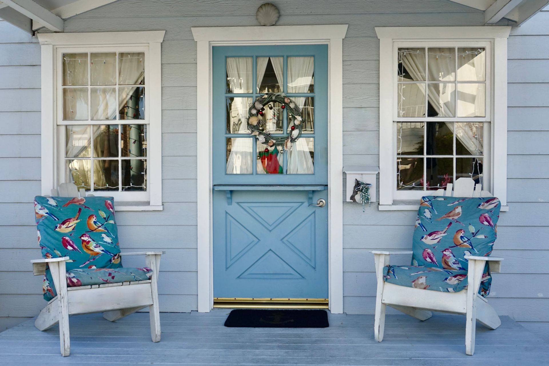 Front porch with blue door, holiday wreath, and two chairs with bird-patterned cushions.