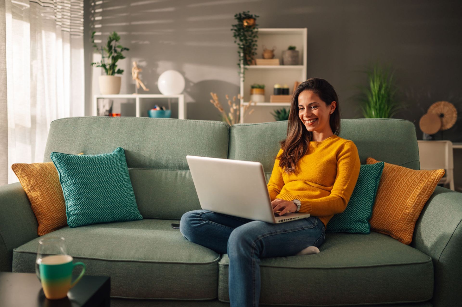 Woman sitting on sofa using a laptop, smiling, with a living room setting in the background.