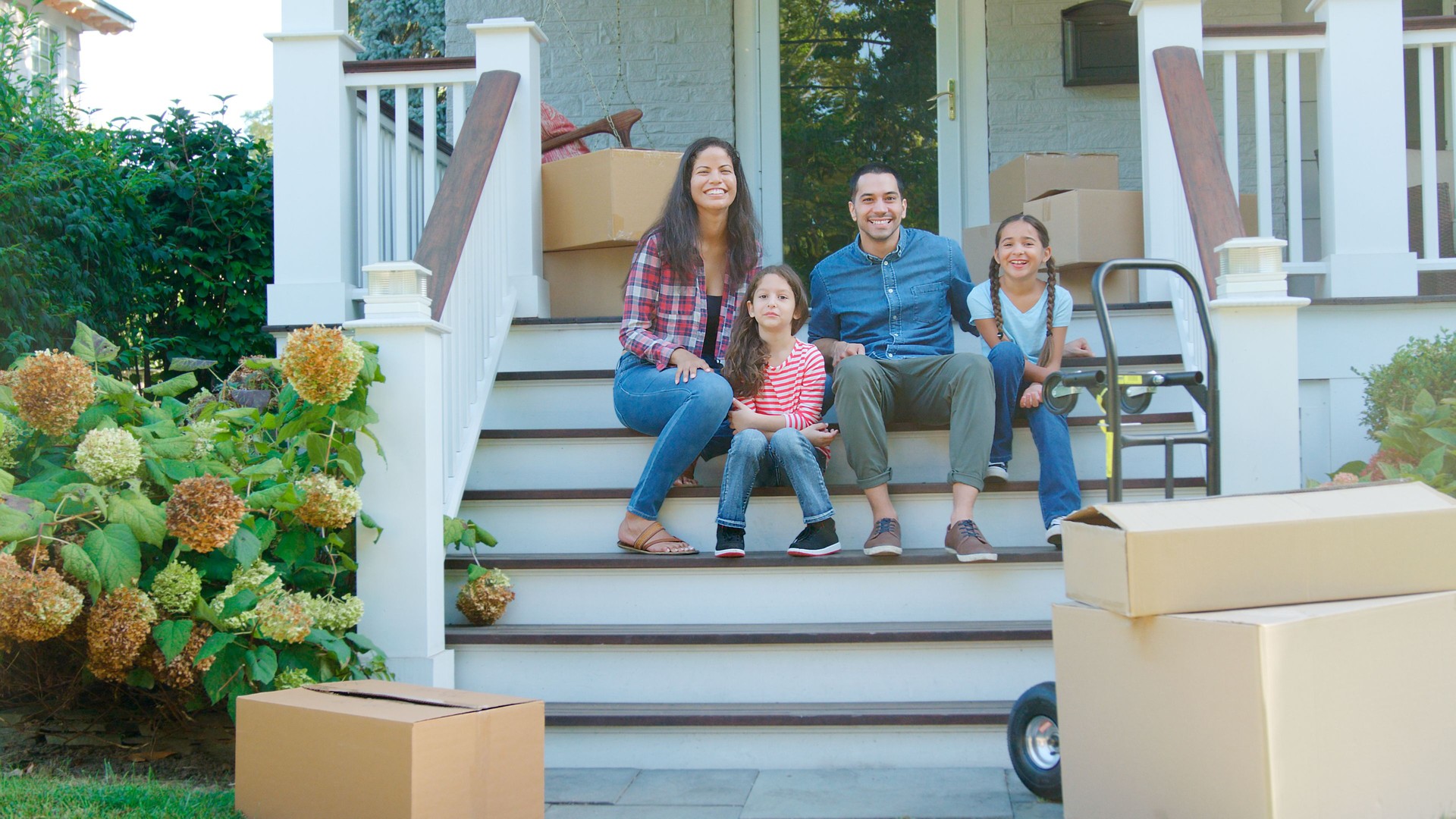 Portrait Of Family Sitting On Steps Of New Home On Moving In Day With Boxes And Trolley