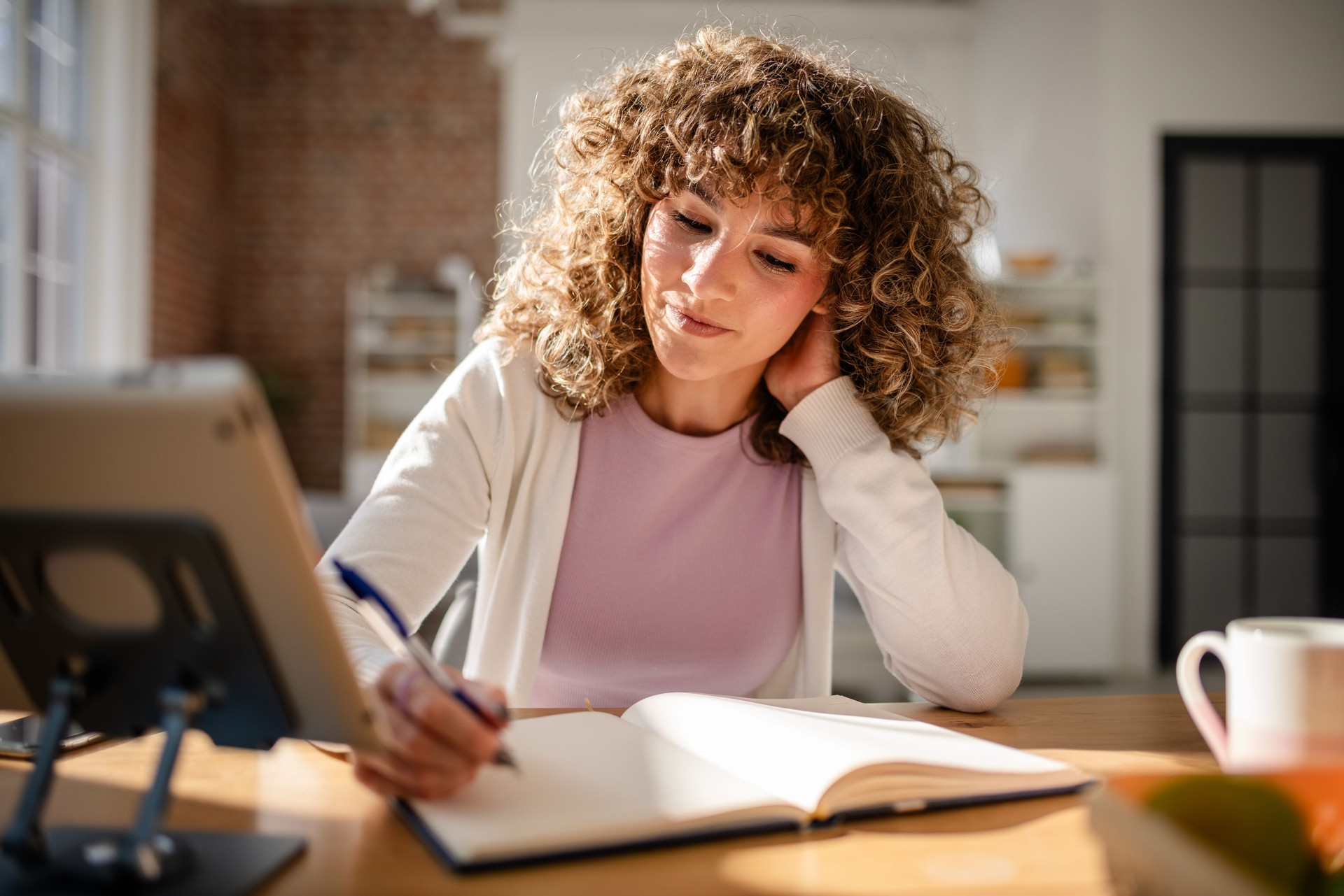 Woman writing in a notebook while working on a tablet in a bright room